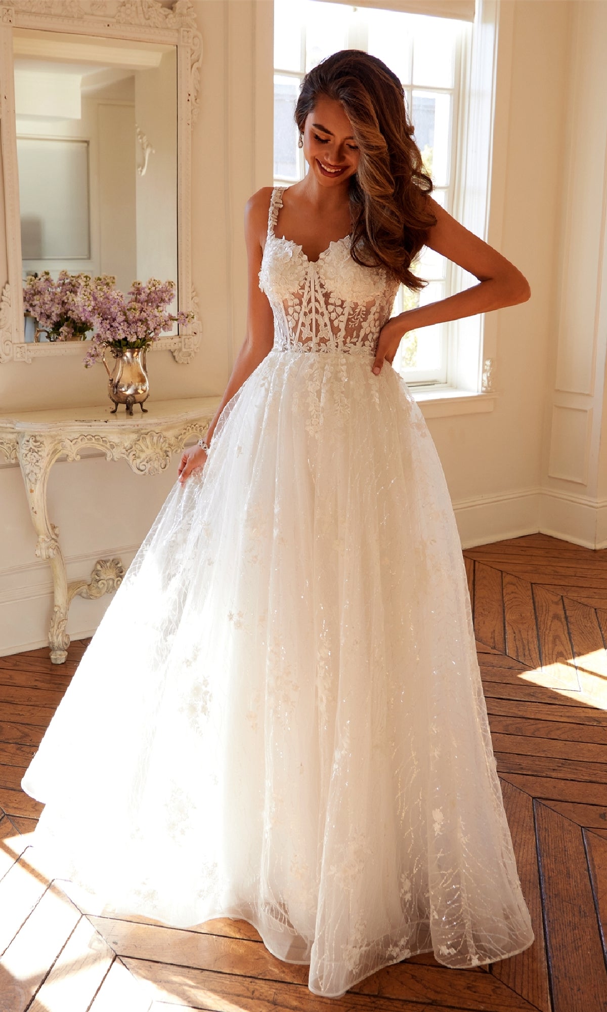 A woman stands in a sunlit room wearing the Juliet JT2495AW long prom dress, featuring an off-white lace corset bodice and full tulle skirt. A vintage vanity with flowers enhances the romantic backdrop.